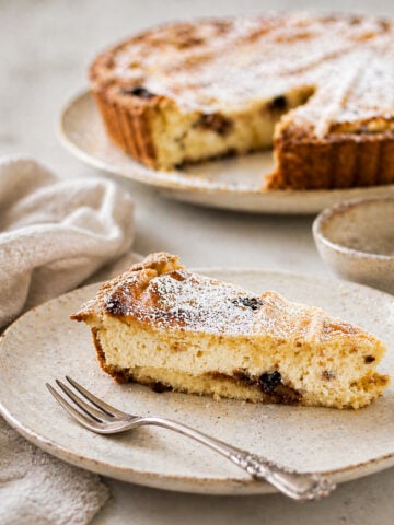 A slice of ricotta and raisin cake, reminiscent of a classic ricotta pie, is topped with powdered sugar and sits on a ceramic plate with a fork. In the background, the rest of the cake waits, partially sliced next to a beige napkin.