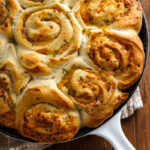 Swirled, golden-brown garlic cheese rolls and cheddar scallion potato buns baked in a white-handled skillet, garnished with herbs, resting on a wooden surface.