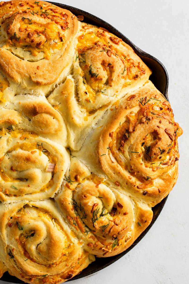 Golden, cheesy garlic herb rolls-reminiscent of cheddar scallion potato buns-baked in a round cast iron skillet, with sprinkles of fresh herbs and melted cheese visible on the soft, fluffy bread swirls.