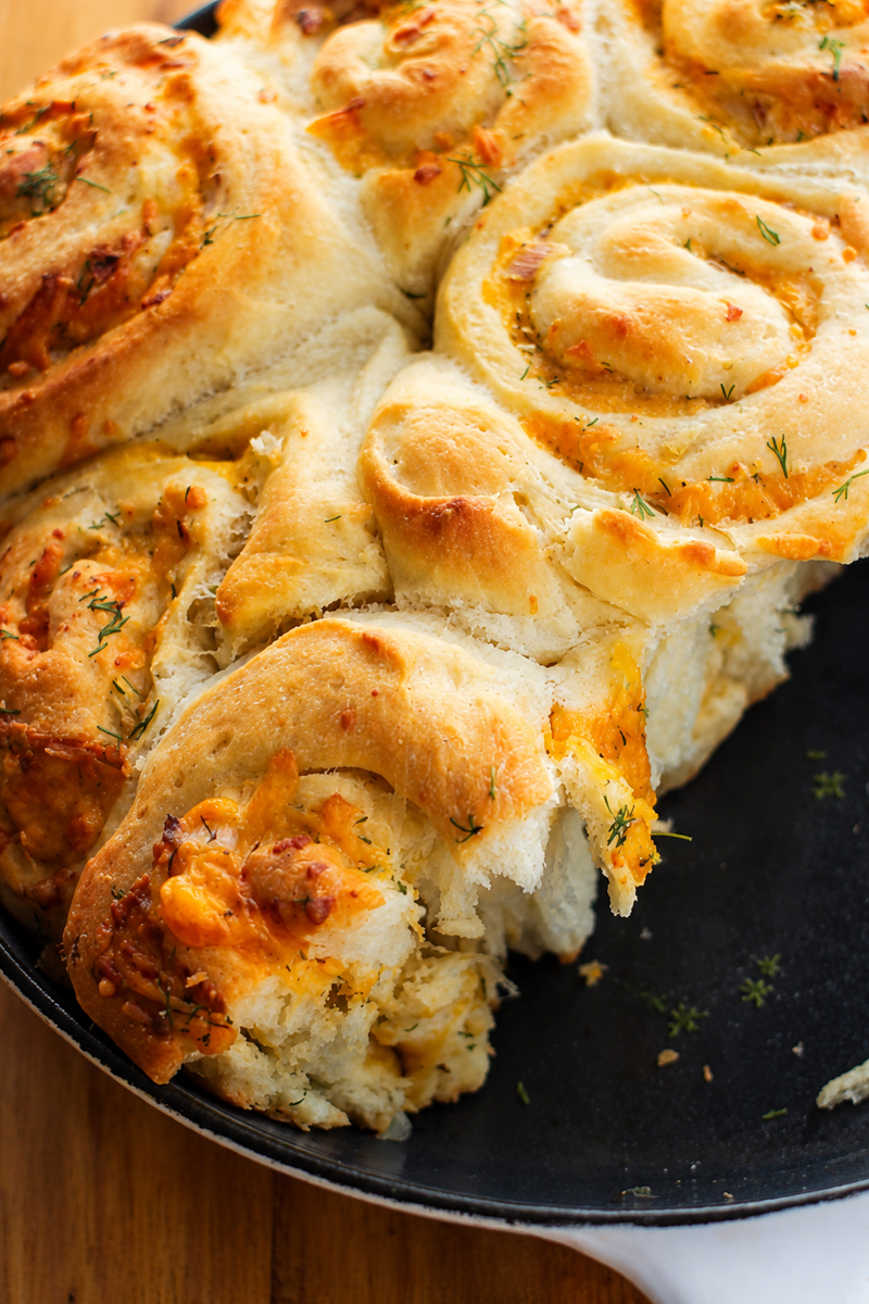 A close-up of cheesy, golden-brown cheddar scallion potato buns in a pull-apart spiral pattern, garnished with fresh herbs, on a black plate.