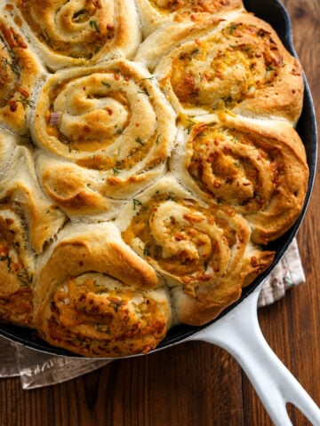 Swirled, golden-brown garlic cheese rolls and cheddar scallion potato buns baked in a white-handled skillet, garnished with herbs, resting on a wooden surface.