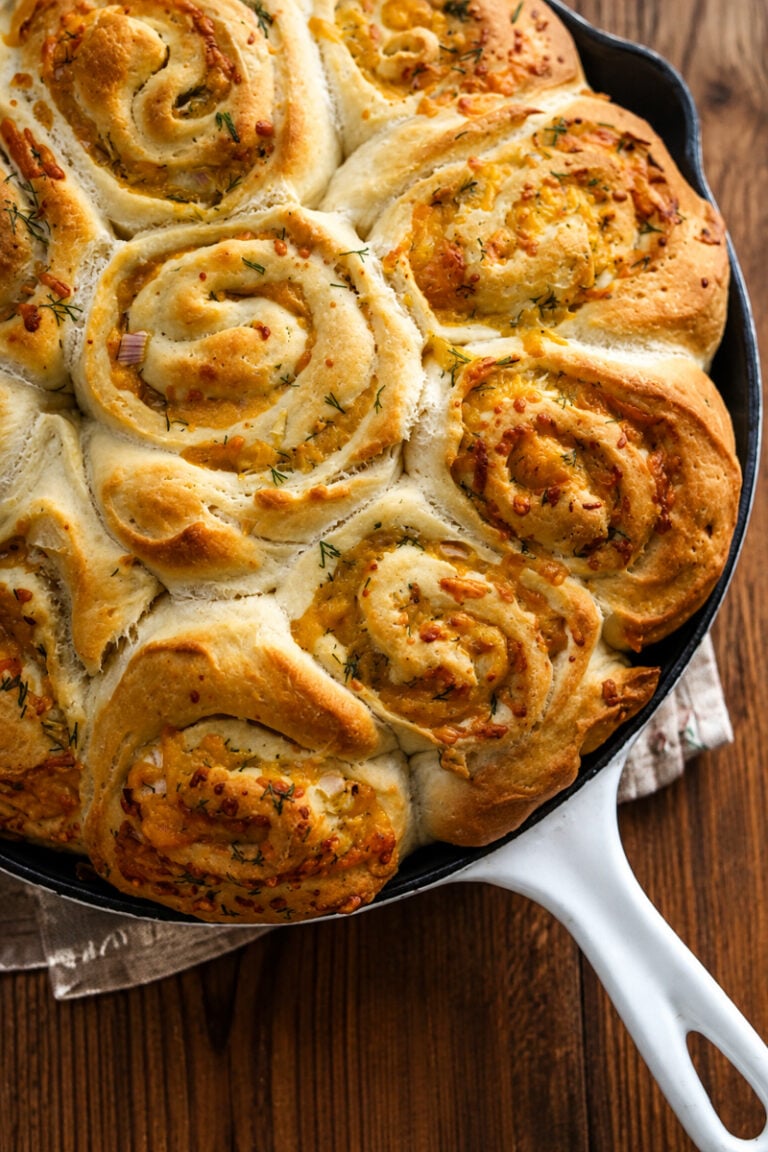 Swirled, golden-brown garlic cheese rolls and cheddar scallion potato buns baked in a white-handled skillet, garnished with herbs, resting on a wooden surface.