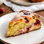 A slice of Raspberry Ricotta Cake sits on a white plate. The cake is golden and moist, with visible raspberries inside and a light dusting of powdered sugar on top. A whole cake and fresh raspberries are in the background.