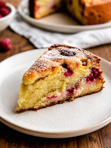 A slice of Raspberry Ricotta Cake sits on a white plate. The cake is golden and moist, with visible raspberries inside and a light dusting of powdered sugar on top. A whole cake and fresh raspberries are in the background.