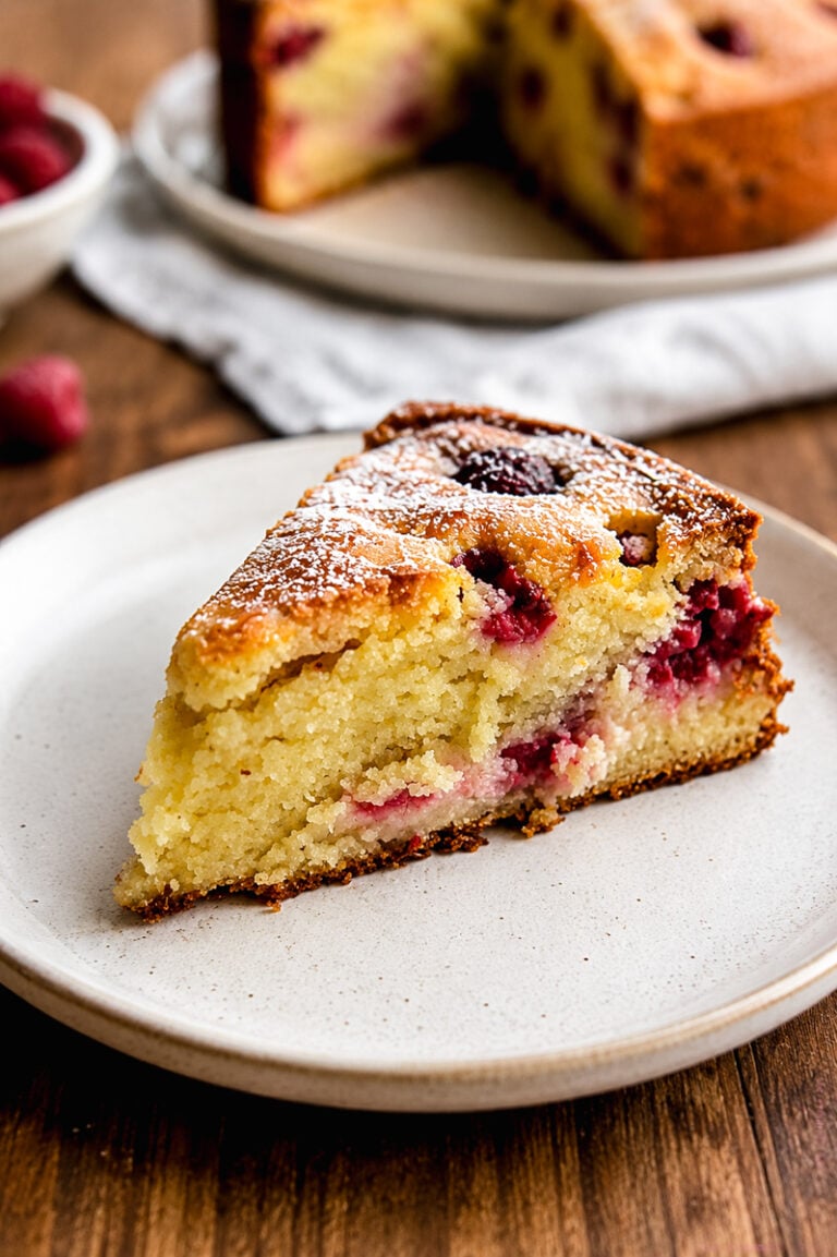 A slice of Raspberry Ricotta Cake sits on a white plate. The cake is golden and moist, with visible raspberries inside and a light dusting of powdered sugar on top. A whole cake and fresh raspberries are in the background.