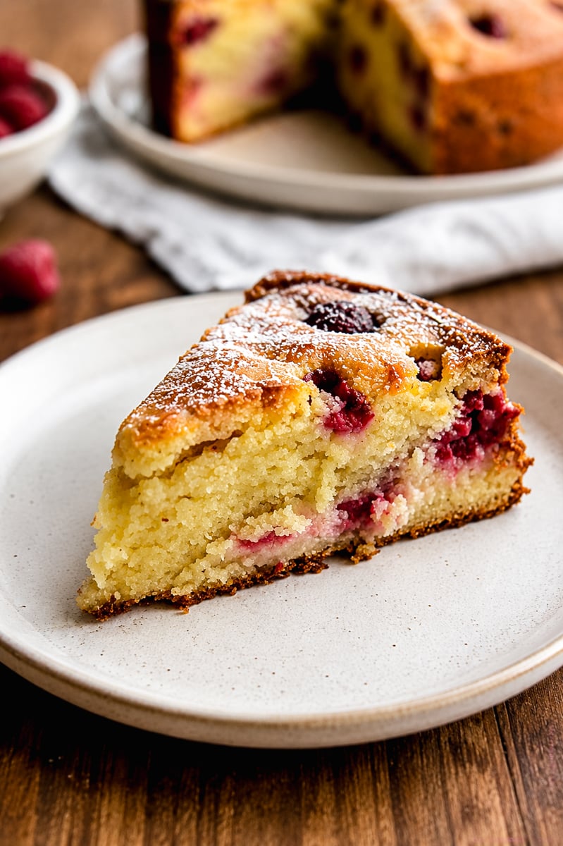 A slice of Raspberry Ricotta Cake sits on a white plate. The cake is golden and moist, with visible raspberries inside and a light dusting of powdered sugar on top. A whole cake and fresh raspberries are in the background.