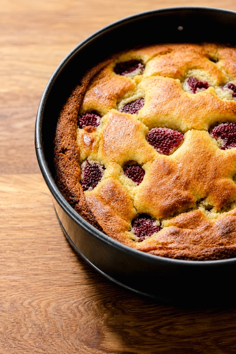 A freshly baked golden Raspberry Ricotta Cake with strawberries on top sits in a round black baking pan on a wooden surface.
