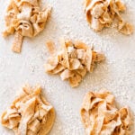 Five small piles of uncooked, homemade pappardelle pasta nests are scattered on a floured surface. The wide pasta ribbons are hand-cut and dusted with flour, ready for cooking.