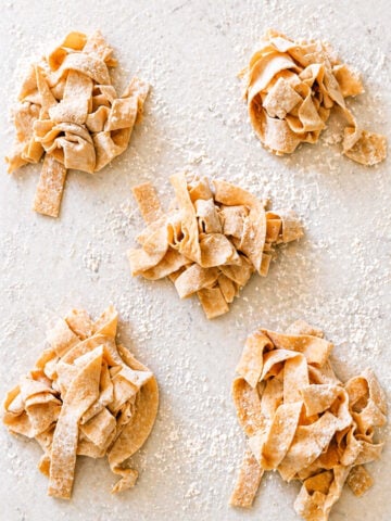 Five small piles of uncooked, homemade pappardelle pasta nests are scattered on a floured surface. The wide pasta ribbons are hand-cut and dusted with flour, ready for cooking.