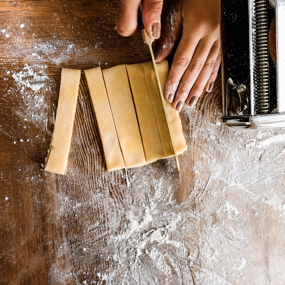 A person cuts rolled-out dough into strips on a floured wooden surface, preparing homemade pappardelle pasta, with a pasta machine visible on the right side.