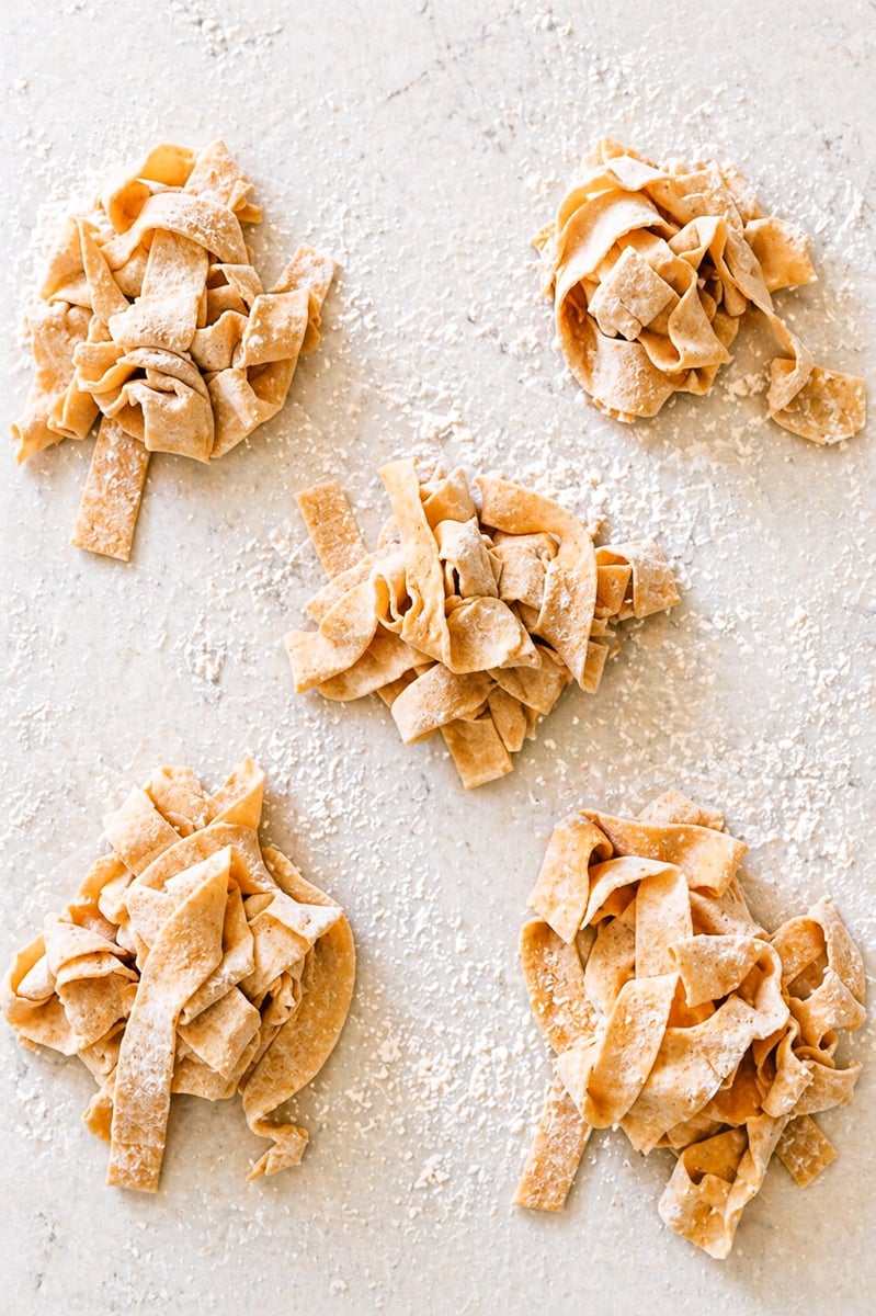 Homemade Pappardelle Five small piles of uncooked, homemade pappardelle pasta nests are scattered on a floured surface. The wide pasta ribbons are hand-cut and dusted with flour, ready for cooking.