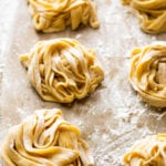 Six nests of fresh, uncooked homemade fettuccine pasta rest on a floured surface, ready for cooking. The pasta strands are lightly dusted with flour to prevent sticking.