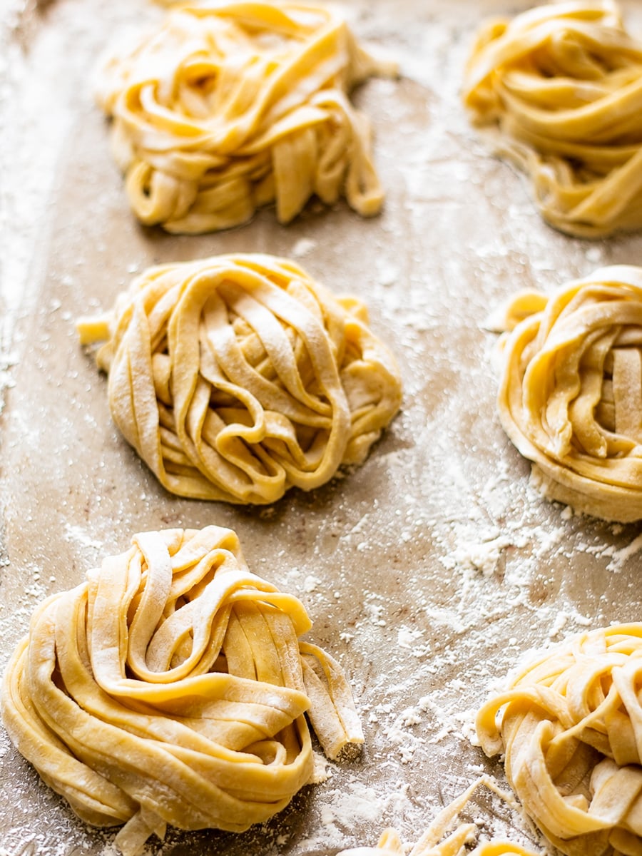 Homemade Fettuccine Six nests of fresh, uncooked homemade fettuccine pasta rest on a floured surface, ready for cooking. The pasta strands are lightly dusted with flour to prevent sticking.
