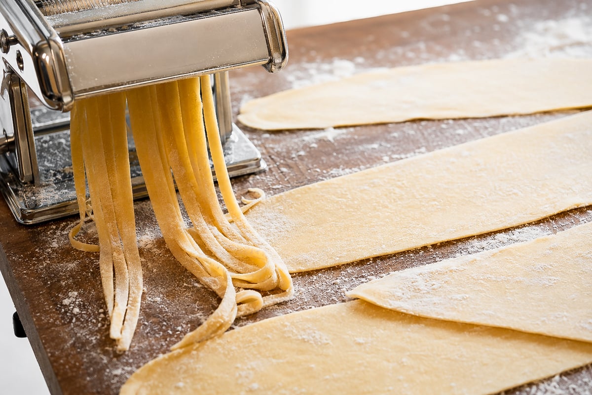 Homemade Fettuccine Homemade fettuccine pasta dough is being rolled and cut into noodles with a metal pasta machine. Sheets of dough and freshly cut homemade fettuccine noodles are arranged on a floured wooden surface.