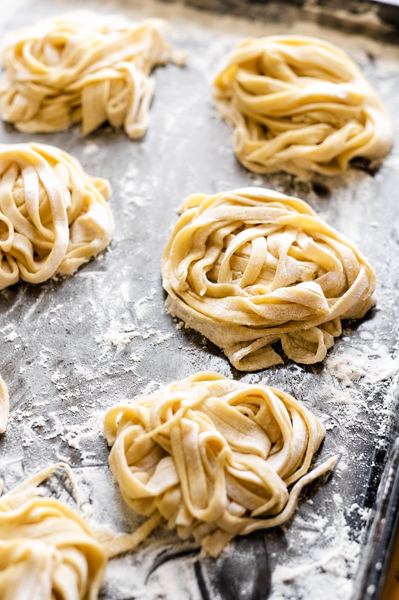 Freshly made nests of homemade fettuccine pasta rest on a floured metal baking sheet, ready to be cooked.