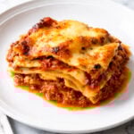 A slice of homemade lasagna, crafted from an authentic lasagna bolognese recipe, features layers of pasta, rich meat sauce, and melted cheese on a white plate, set on a marble surface.