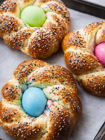 Three braided Italian Easter breads topped with colorful sprinkles, each with a brightly dyed egg (green, blue, pink) in the center, sit on a parchment-lined baking tray.