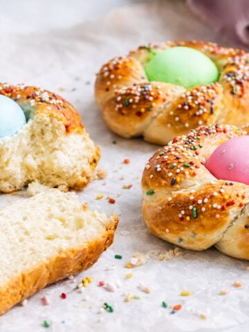 Three braided Italian Easter breads, each with a colored egg in the center and topped with rainbow sprinkles. One bread is partially eaten, a piece torn off, and sprinkles are scattered on the parchment paper.