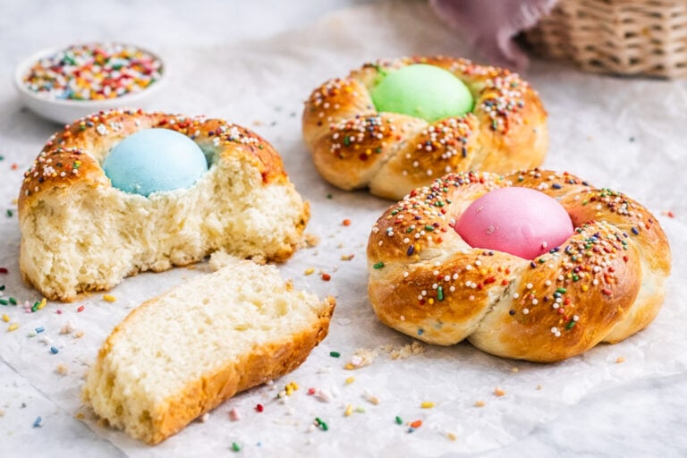 Three braided Italian Easter breads, each with a colored egg in the center and topped with rainbow sprinkles. One bread is partially eaten, a piece torn off, and sprinkles are scattered on the parchment paper.