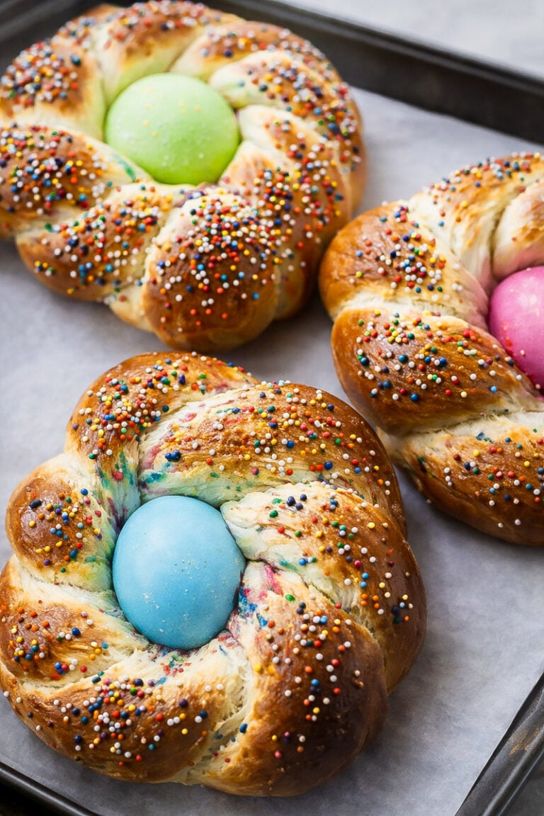 Three braided Italian Easter breads topped with colorful sprinkles, each with a brightly dyed egg (green, blue, pink) in the center, sit on a parchment-lined baking tray.