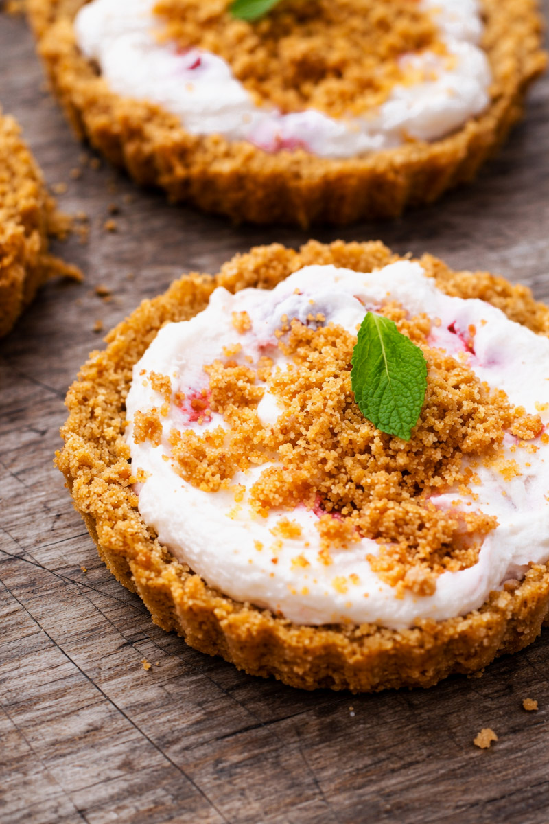 A close-up of a No-Bake Strawberry and Mascarpone Cheesecake with a crumbly graham cracker crust, creamy filling, and a sprinkling of crumbs on top, garnished with a small mint leaf, served on a rustic wooden surface.