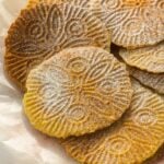 A close-up of several gluten-free pizzelle cookies with intricate floral patterns, dusted with powdered sugar, arranged on a plate lined with parchment paper.