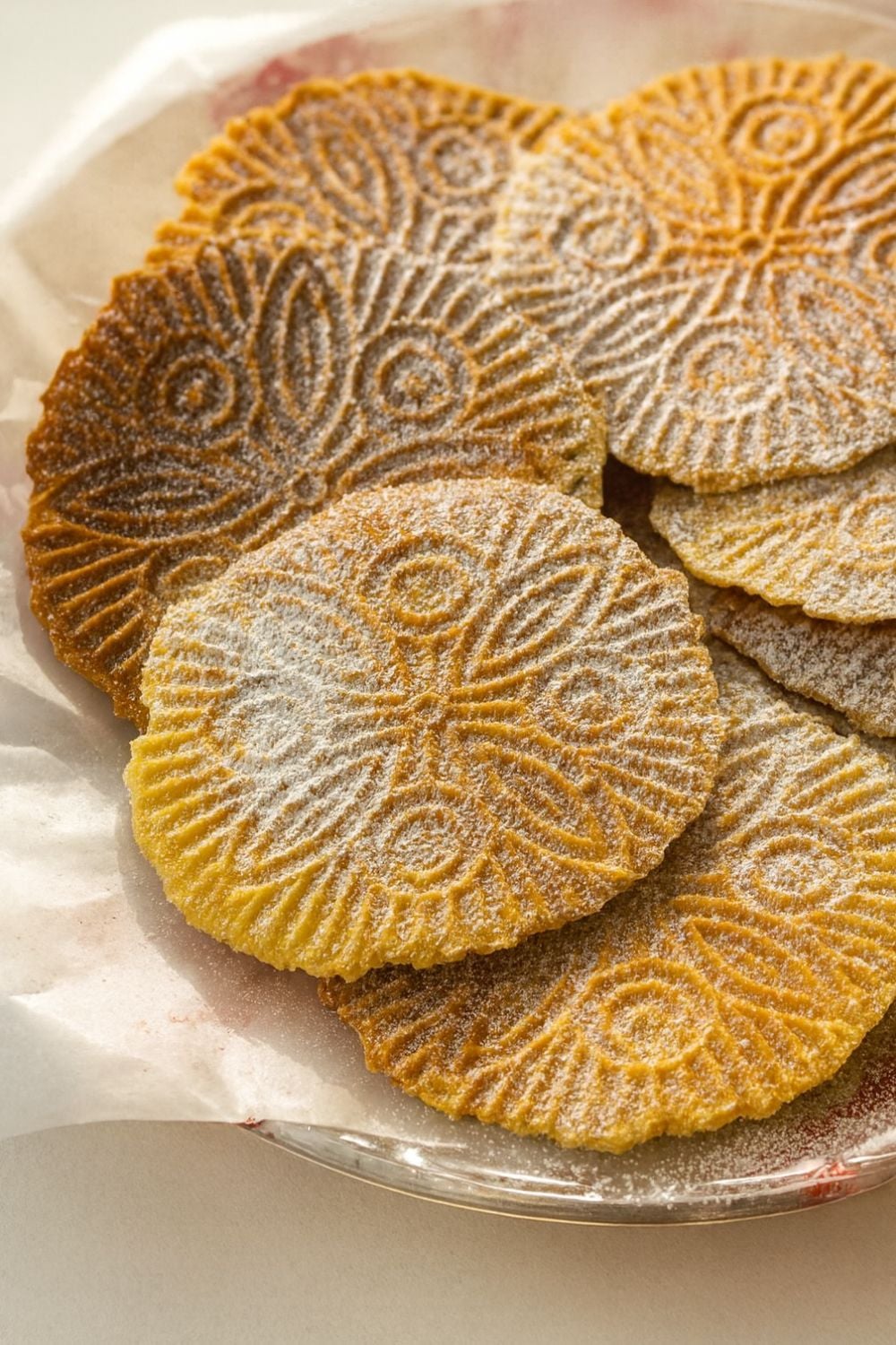 A close-up of several gluten-free pizzelle cookies with intricate floral patterns, dusted with powdered sugar, arranged on a plate lined with parchment paper.