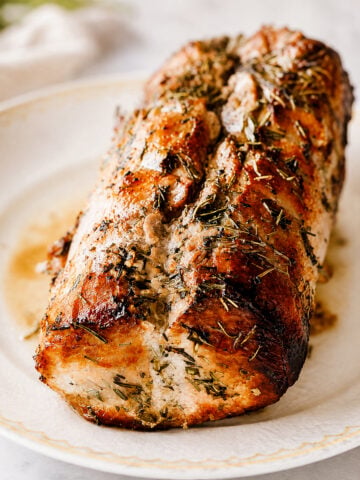 A roasted pork loin-Roast Pork Loin-Arrosto di Maiale-garnished with herbs sits on a white plate, showing a golden-brown, crispy exterior and juicy, tender interior.
