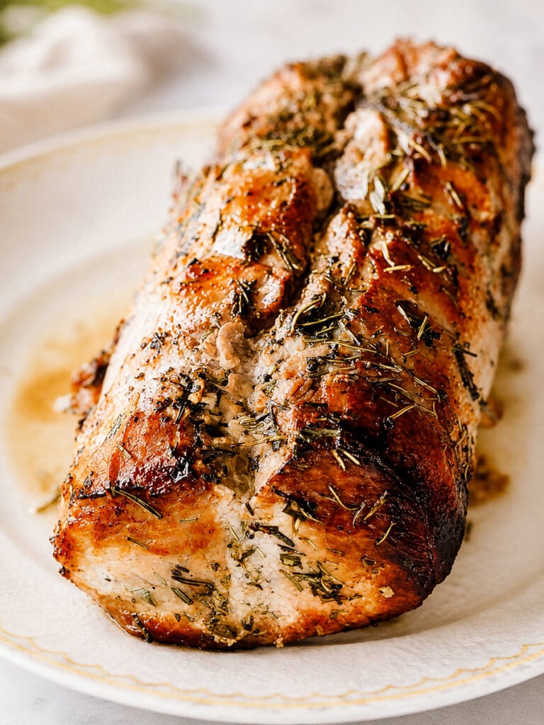 A roasted pork loin—Roast Pork Loin-Arrosto di Maiale—garnished with herbs sits on a white plate, showing a golden-brown, crispy exterior and juicy, tender interior.