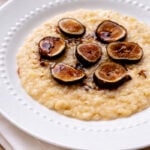 A white plate with creamy Risotto with Figs and Prosecco-Risotto con Fichi e Prosecco, topped with sliced figs and a drizzle of dark sauce, sits on a white napkin, accented by fresh herbs in the background.