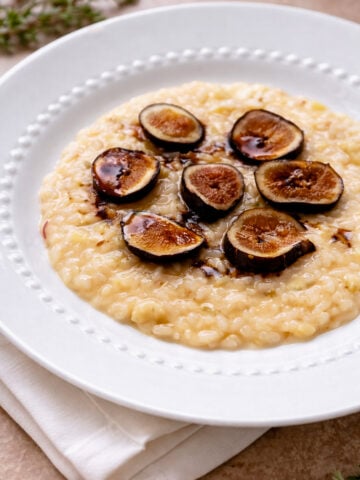 A white plate with creamy Risotto with Figs and Prosecco-Risotto con Fichi e Prosecco, topped with sliced figs and a drizzle of dark sauce, sits on a white napkin, accented by fresh herbs in the background.