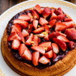 A Strawberry Sponge Cake topped with strawberry jam, fresh sliced strawberries, and lemon zest sits on a decorative white plate; a bowl of strawberries and a lemon are in the background.