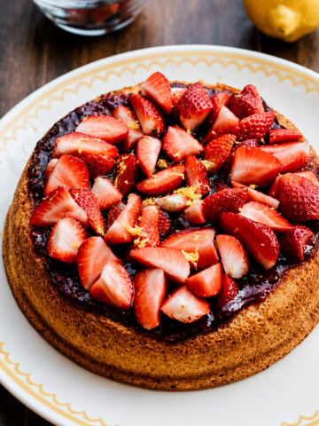 A Strawberry Sponge Cake topped with strawberry jam, fresh sliced strawberries, and lemon zest sits on a decorative white plate; a bowl of strawberries and a lemon are in the background.