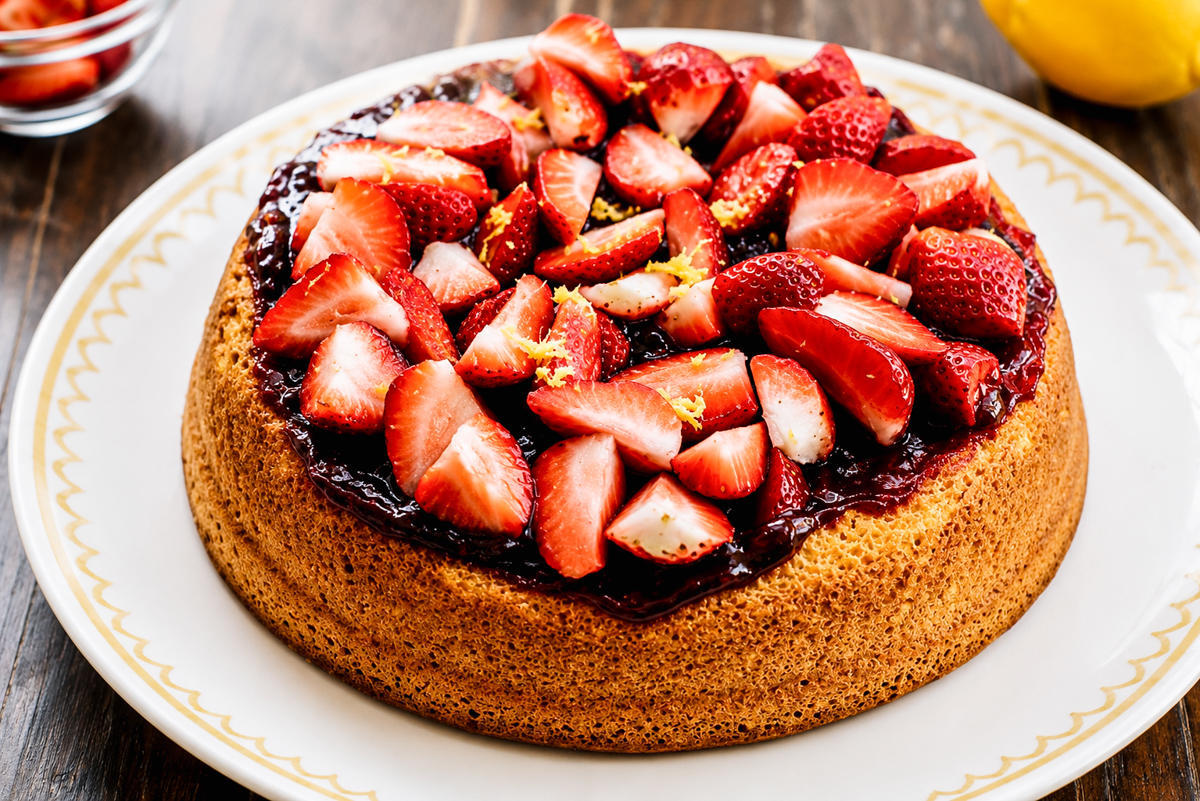 A round Strawberry Sponge Cake topped with a layer of strawberry jam and fresh sliced strawberries, served on a white plate with a decorative gold rim. A lemon and a bowl of strawberries are in the background.