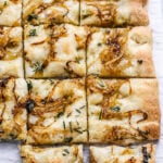 A close-up of golden, square pieces of Rosemary and Caramelized Onion Focaccia topped with caramelized onions and chopped herbs, arranged on parchment paper. One piece is slightly pulled away from the rest.