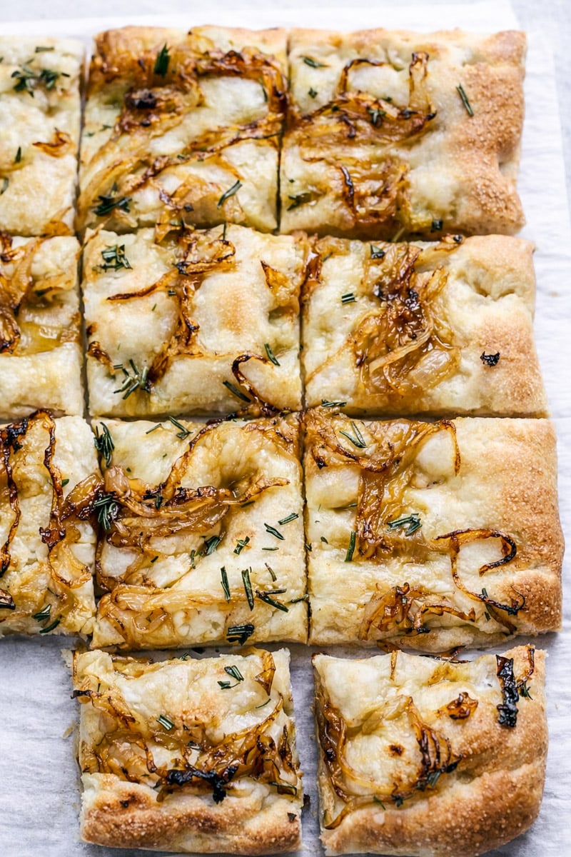 A close-up of golden, square pieces of Rosemary and Caramelized Onion Focaccia topped with caramelized onions and chopped herbs, arranged on parchment paper. One piece is slightly pulled away from the rest.