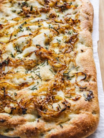 A Rosemary and Caramelized Onion Focaccia sits on parchment paper, topped with caramelized onions, melted cheese, and chopped rosemary, showcasing a golden, crispy crust and airy texture.