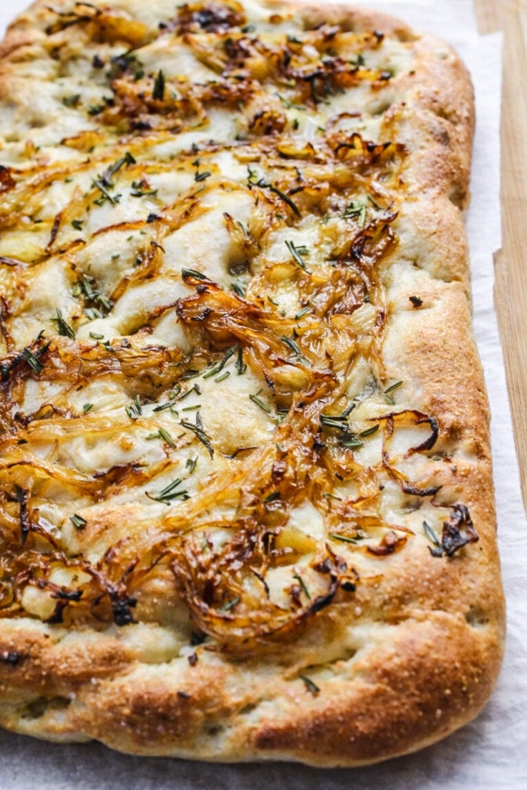 A Rosemary and Caramelized Onion Focaccia sits on parchment paper, topped with caramelized onions, melted cheese, and chopped rosemary, showcasing a golden, crispy crust and airy texture.