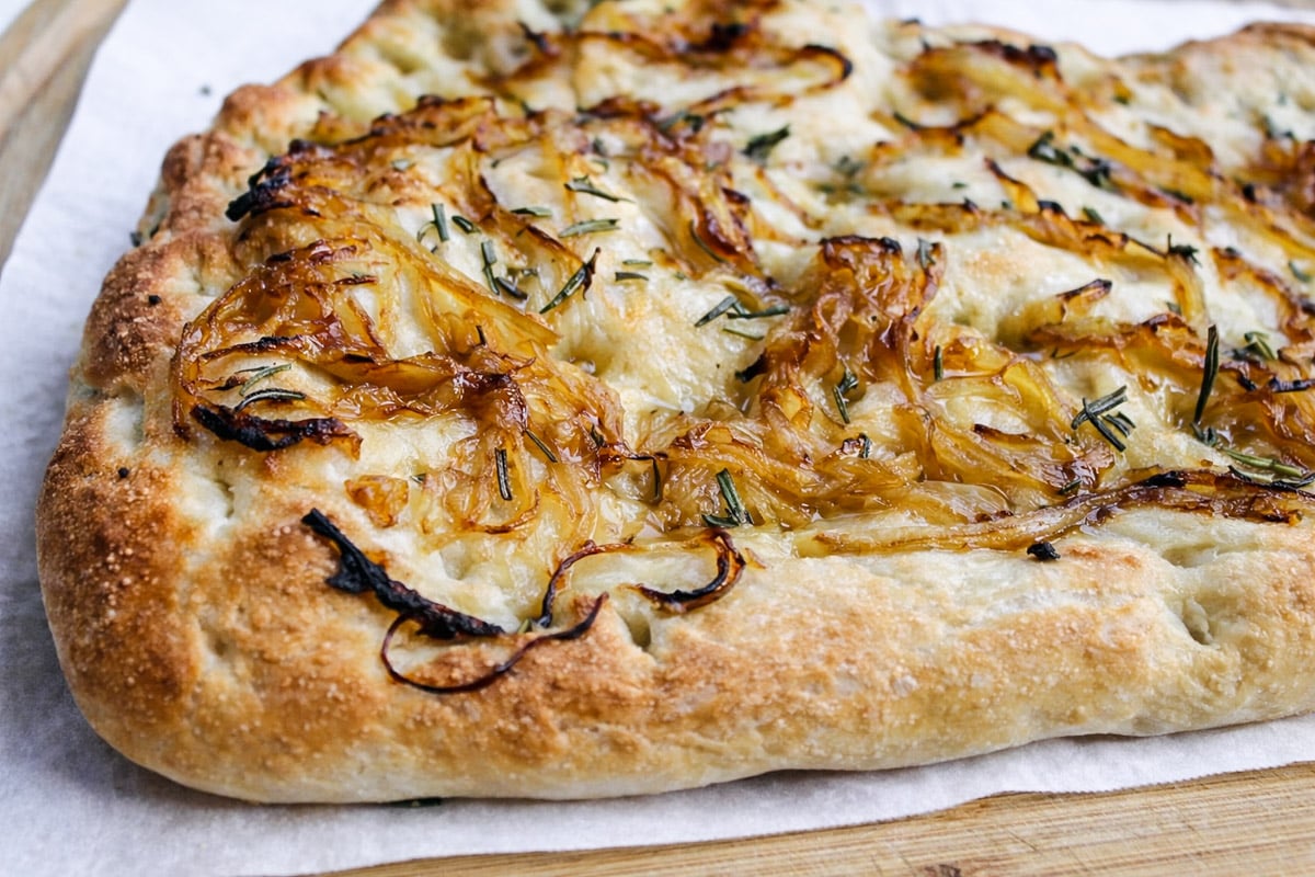 A close-up of Rosemary and Caramelized Onion Focaccia, its golden-brown crust topped with sweet onions and fresh rosemary, resting on parchment paper atop a wooden board.
