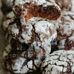 A stack of Chocolate Crackle Cookies dusted with powdered sugar, with one cookie on top showing a bite taken out, revealing a soft, chocolatey interior.