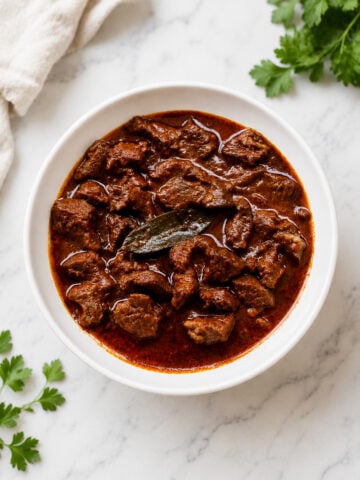 A white bowl filled with spicy beef curry, reminiscent of Hungarian Goulash Soup-Gulyásleves, garnished with a bay leaf and placed on a white marble surface with fresh cilantro leaves nearby.