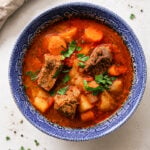 A blue-patterned bowl filled with hearty Hungarian Goulash Soup-Gulyásleves containing chunks of beef, potatoes, carrots, and fresh herbs, set on a light background with a beige napkin nearby.