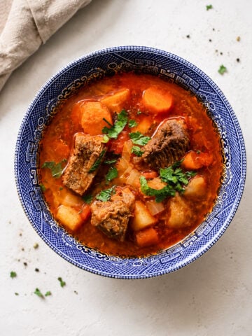 A blue-patterned bowl filled with hearty Hungarian Goulash Soup-Gulyásleves containing chunks of beef, potatoes, carrots, and fresh herbs, set on a light background with a beige napkin nearby.