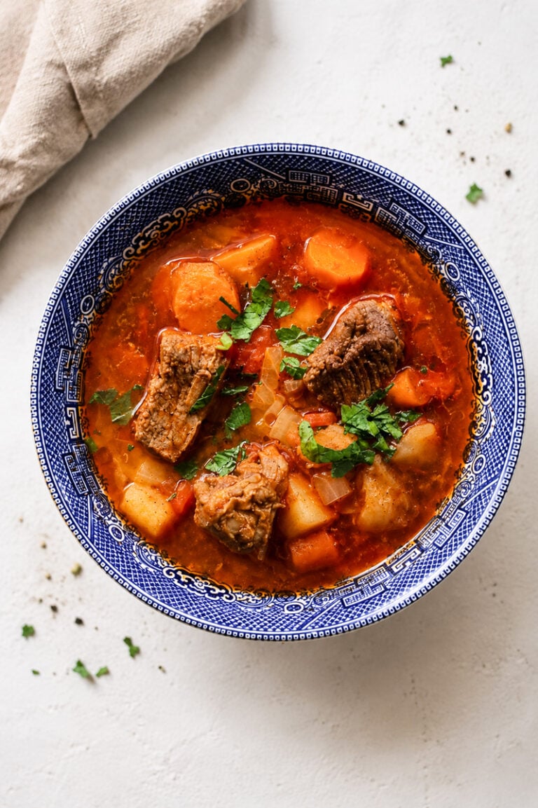 A blue-patterned bowl filled with hearty Hungarian Goulash Soup-Gulyásleves containing chunks of beef, potatoes, carrots, and fresh herbs, set on a light background with a beige napkin nearby.