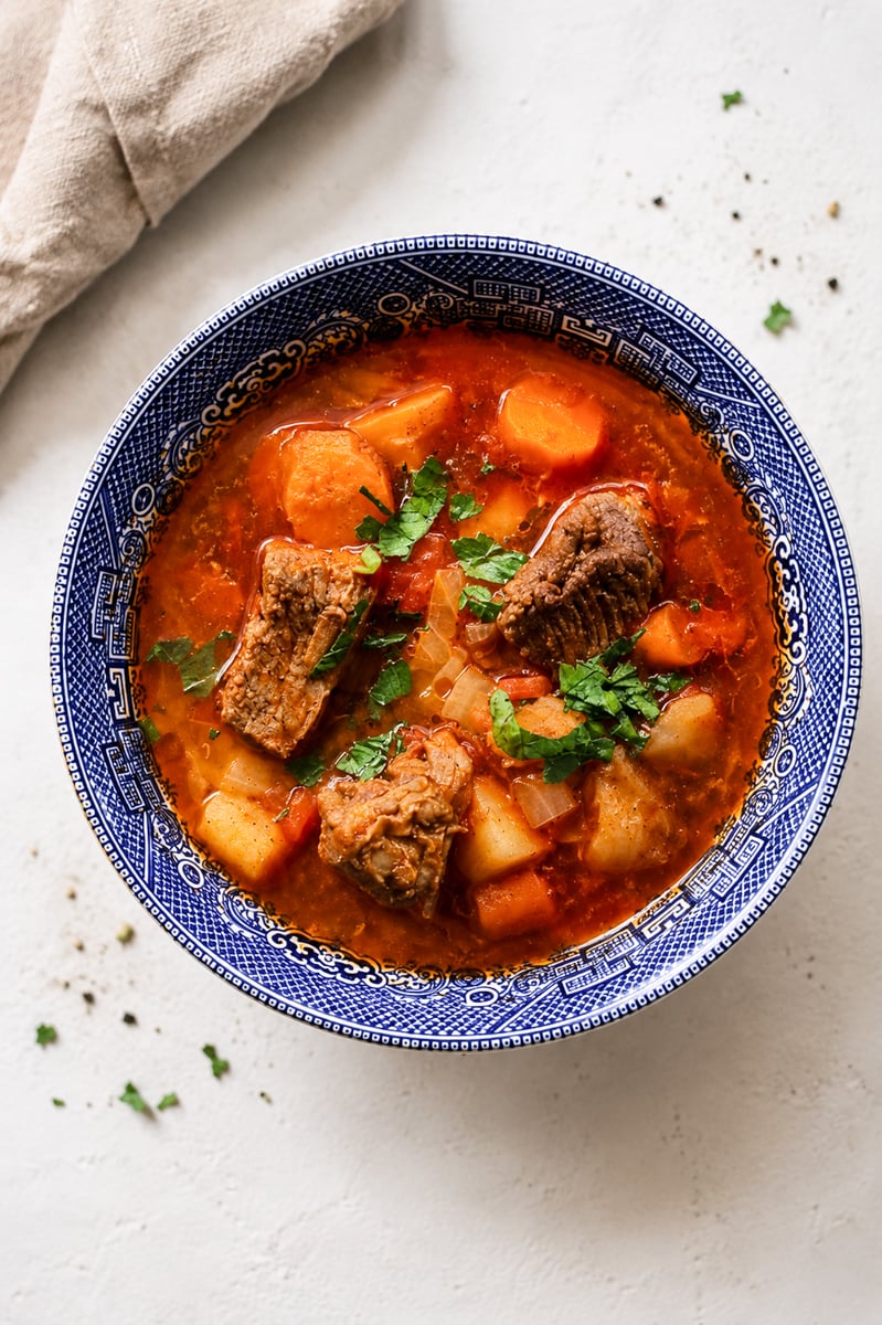 A blue-patterned bowl filled with hearty Hungarian Goulash Soup-Gulyásleves containing chunks of beef, potatoes, carrots, and fresh herbs, set on a light background with a beige napkin nearby.