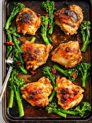 A baking sheet with six golden-brown Sheet Pan Honey Balsamic Chicken Thighs, surrounded by fresh green broccolini and sun-dried tomato pieces, with a fork resting on the tray.