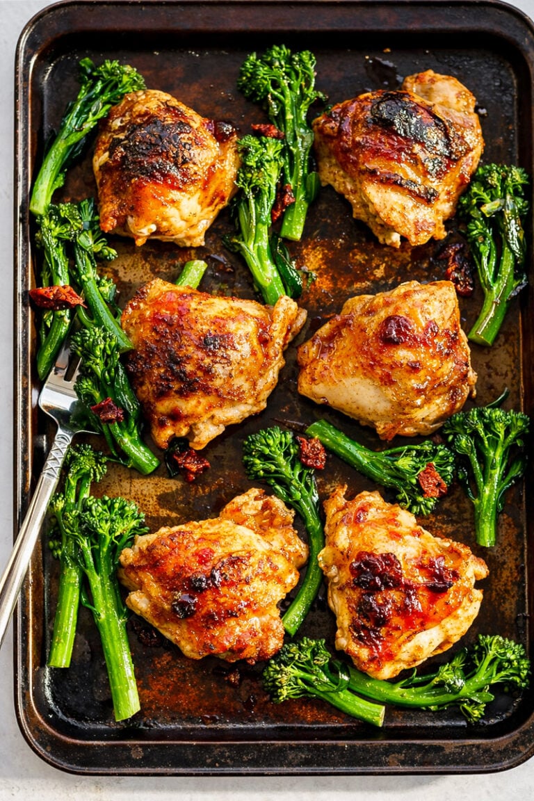 A baking sheet with six golden-brown Sheet Pan Honey Balsamic Chicken Thighs, surrounded by fresh green broccolini and sun-dried tomato pieces, with a fork resting on the tray.