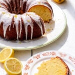 A lemon curd cake bundt with white glaze sits on a plate, with a slice served on a floral-patterned dish. Fresh lemons, some halved, are scattered nearby on a white surface.