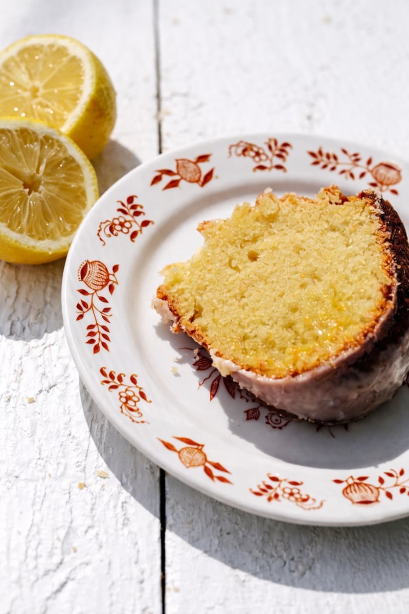 This Lemon Bundt Cake with Lemon Curd Filling is sure to brighten any party or special event! It is bursting with lemon flavor from the lemon zest, homemade lemon curd and fresh lemon glaze. #baking #lemon #bundtcake #lemoncurd #dessert A slice of lemon curd cake with a golden crust sits on a white floral-patterned plate, next to two lemon halves, on a rustic white wooden surface.