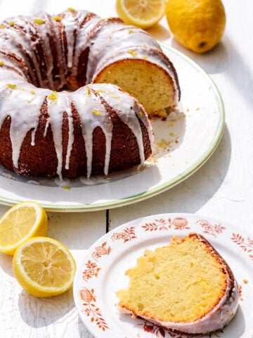 A lemon curd cake bundt with white glaze sits on a plate, with a slice served on a floral-patterned dish. Fresh lemons, some halved, are scattered nearby on a white surface.